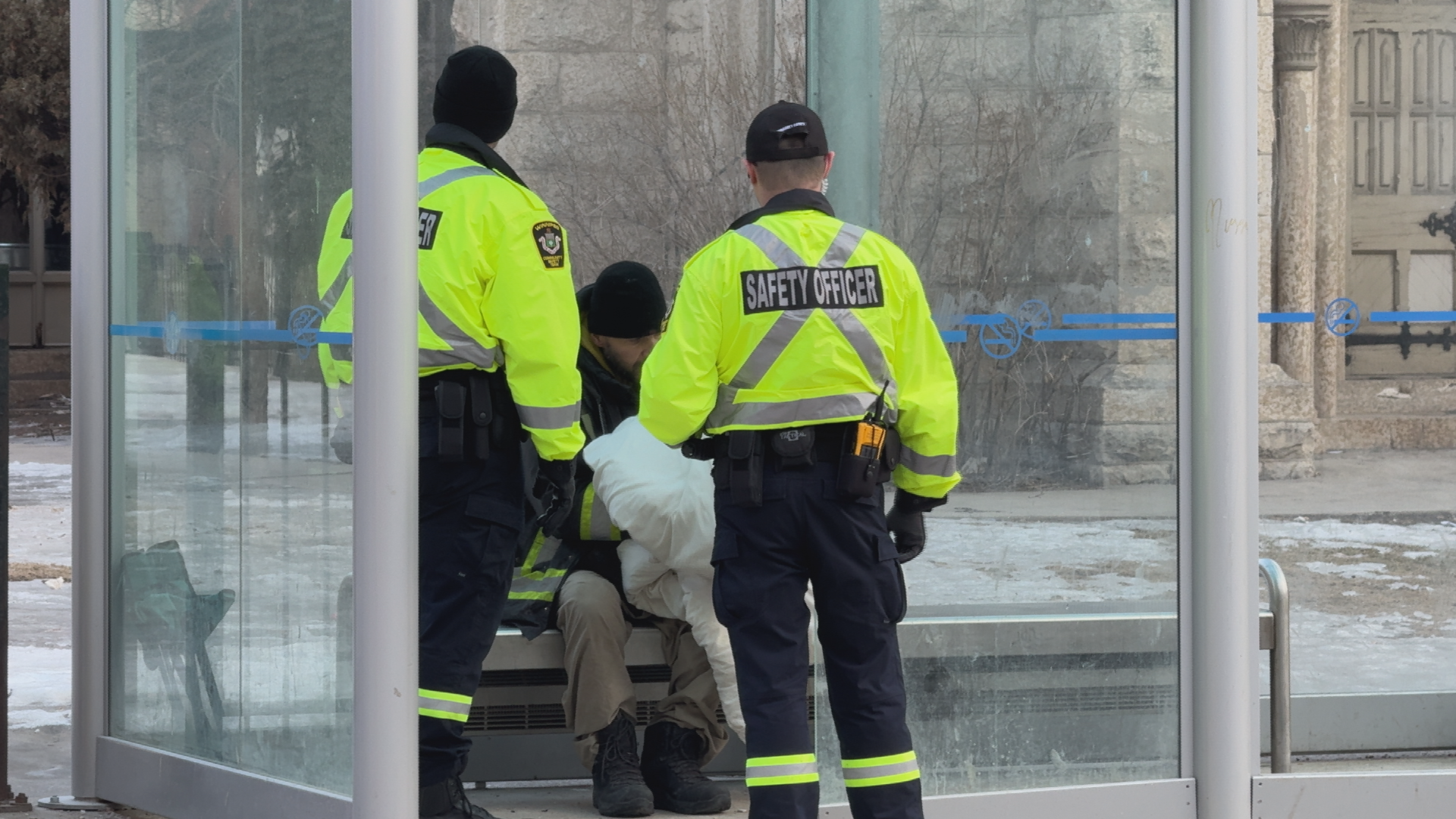 Photo of two transit security in bright yellow jackets speaking to a person in a bus shelter