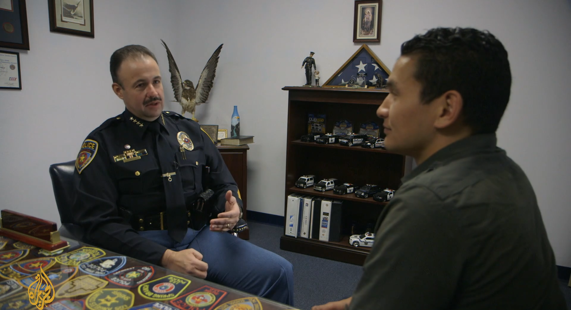 Screenshot from documentary that shows a police officer sitting in his office and Wab Kinew sitting opposite him. There are police badges and toy cars all over the office.